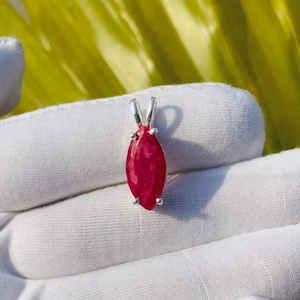 May include: A ruby pendant with a silver bail. The pendant features a marquise-cut, deep red gemstone. The pendant is held by a gloved hand against a background of green leaves.