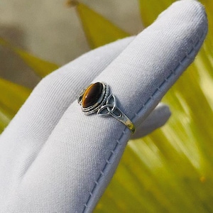 May include: A silver ring with an oval tiger's eye stone. The ring has a Celtic knot design on the sides of the stone. The ring is displayed on a white glove against a blurred green and yellow background.