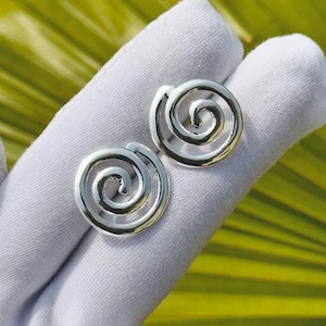 May include: A pair of silver-colored spiral stud earrings. The earrings are round with a raised spiral design. The earrings are displayed on a white surface against a background of green leaves.