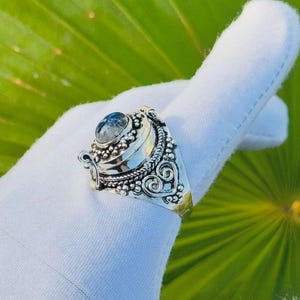 May include: A silver ring with an oval, light blue gemstone. The ring has an ornate design with small beaded details and swirling patterns. The ring is shown on a white surface against a green background.