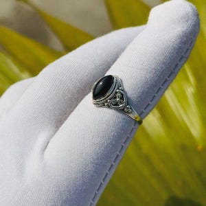 Op de afbeelding: Een zilveren ring met een ovale, zwarte edelsteen. De ring heeft een sierlijk ontwerp rond de steen en op de band. De ring wordt gepresenteerd op een wit oppervlak, met een vage groen-gele achtergrond.