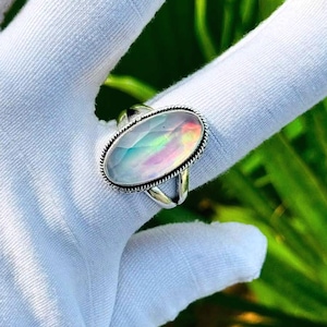 May include: An oval-shaped ring featuring a faceted, iridescent stone with a milky, translucent appearance. The stone is set in a polished silver-toned metal band with a decorative twisted rope texture along the outer edge of the setting.