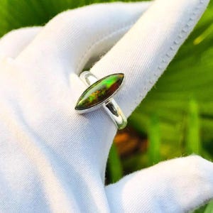 May include: A silver ring featuring a marquise-cut iridescent gemstone with shifting green, orange, and red hues. The stone is set in a polished metal band that encircles the finger, showcasing a vibrant play of color within the gem.
