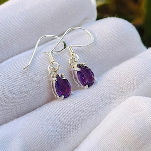 Könnte beinhalten: Silberne Ohrhänger mit tropfenförmigen, tiefvioletten Edelsteinen. Die Ohrringe werden auf einer weißen Oberfläche präsentiert, mit einem verschwommenen Hintergrund aus grünem Laub und einer roten Blume.