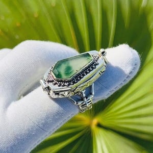 Op de afbeelding: Een zilveren ring met een lichtgroene, smaragdgeslepen edelsteen. De ring heeft een gedetailleerd, sierlijk ontwerp met kleine, donkere accenten rond de edelsteen. De ring wordt getoond tegen een achtergrond van groene bladeren.