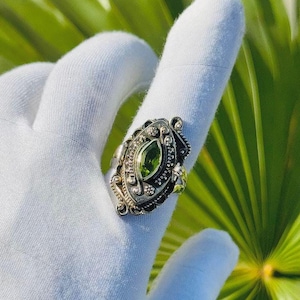 May include: An ornate silver ring featuring a large, oval-cut peridot gemstone. The ring has intricate filigree detailing and small accent stones. The peridot is a vibrant green color, set against the silver band. The ring is displayed on a white surface.