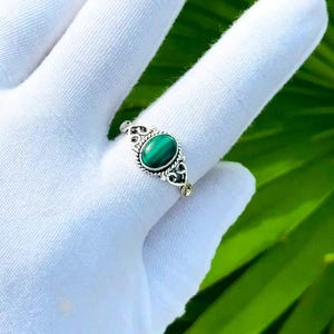 May include: A silver ring featuring an oval-shaped green malachite gemstone set in a decorative metal band with intricate twisted wire and scrollwork detailing. The stone is smooth and polished, displaying deep green banding patterns.