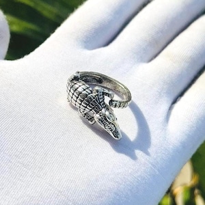 Op de afbeelding: Een zilverkleurige metalen ring in de vorm van een alligator. Het ontwerp heeft een getextureerde, geschubde rug en een gedetailleerde kop die om de vinger wikkelt. Het metaal heeft een donkere, geoxideerde afwerking in de groeven.
