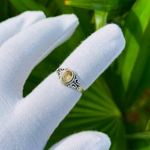 Op de afbeelding: Een zilveren ring met een ovaalvormige gele edelsteen in een decoratieve metalen band met ingewikkeld krulwerk en kraaldetails, gedragen aan een vinger bedekt met een witte stoffen handschoen tegen een achtergrond van groene palmbladeren.