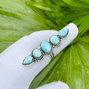 Puede incluir: Un anillo de plata con cinco piedras preciosas de color azul claro, de forma ovalada y de lágrima. El anillo se muestra sobre un fondo blanco, con hojas verdes visibles. Las piedras están dispuestas en línea.