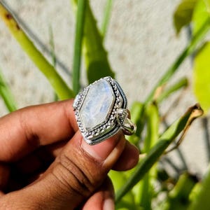 Könnte beinhalten: Ein Silberring mit einem großen, facettierten, hellfarbigen Edelstein in einer einzigartigen geometrischen Form. Der Ring hat aufwendige Details um den Stein und am Band, die eine Vintage- oder handgefertigte Ästhetik zeigen. Der Ring wird vor einem Hintergrund aus grünem Laub gehalten.