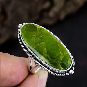 May include: An oval-shaped ring featuring a large, smooth, lime green stone with subtle veining. The stone is set in a polished silver-toned metal frame with a decorative twisted rope border and small bead accents along the outer edge of the setting.