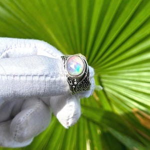 May include: A silver ring featuring a round, faceted, iridescent stone with a play of color. The band has a dark, oxidized finish with intricate, raised, swirling patterns that wrap around the finger. The metalwork has a textured, relief design.