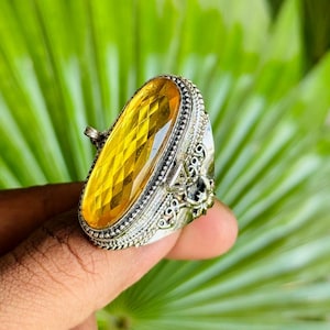 May include: A silver ring with an oval, faceted yellow gemstone. The ring has intricate detailing around the stone and on the band. The gemstone is a bright, sunny yellow color. The ring is held up by a person's finger.