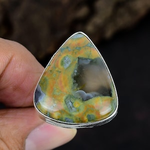 May include: A triangular ring featuring a polished, multi-colored stone with swirling patterns of mustard yellow, sage green, and translucent gray. The stone is set in a smooth, silver-toned metal bezel that follows the triangular shape of the gem.