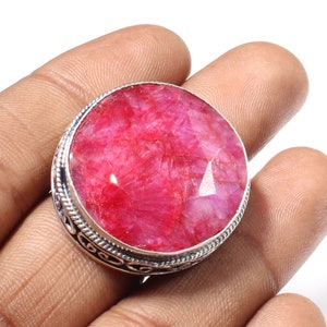 May include: A silver ring with a large, faceted, red ruby gemstone. The ring has an ornate, silver band with a decorative design.