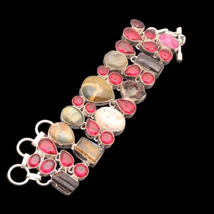 Könnte beinhalten: Ein silbernes Armband mit einer Vielzahl von Edelsteinen in Rot-, Braun-, Weiß- und Grüntönen. Das Armband zeigt eine Mischung aus Formen, darunter Tropfen, Ovale und unregelmäßige Formen, vor einem schwarzen Hintergrund.