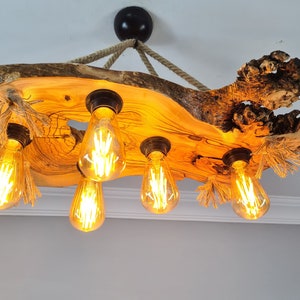 Può includere: Lampadario rustico in legno con sei lampadine in stile Edison. L'apparecchio è sospeso al soffitto con una corda. Il legno ha una forma naturale e irregolare e un colore caldo e dorato, con venature e nodi visibili. Le lampadine emettono un bagliore caldo e invitante.