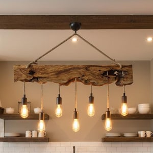 Puede incluir: Isla de cocina rústica con encimera de madera y dos taburetes de bar de cuero marrón. Encima, una lámpara única hecha de una viga de madera cuelga con seis bombillas estilo Edison. La cocina presenta azulejos blancos y estantes de madera.