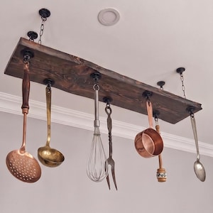 May include: A rectangular dark stained wooden board suspended from the ceiling by black chains and hardware. Hanging from the board are various kitchen utensils including a copper skimmer, a brass ladle, a metal whisk, a fork, a copper pot, and a spoon.