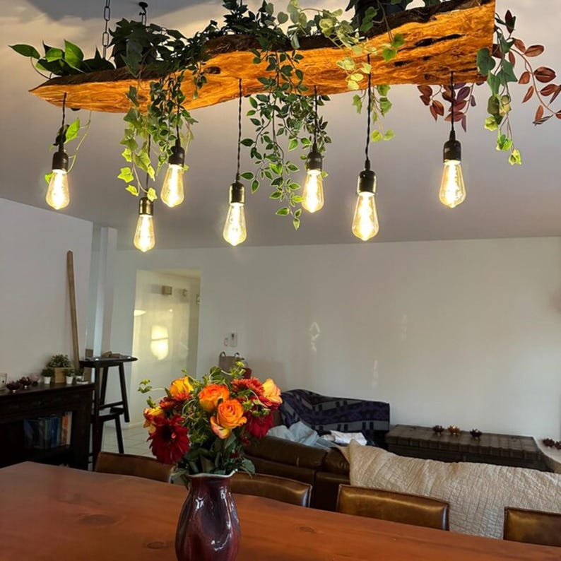 May include: A rustic wooden light fixture with six Edison bulbs hanging from it. The fixture is decorated with green plants and vines. The fixture is hanging over a wooden table with a vase of flowers on it. The table is in a living room with a couch and a coffee table in the background.