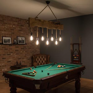 May include: A billiards room featuring a green felt pool table with a wooden frame and legs. Above the table hangs a rustic chandelier with exposed light bulbs. The room has a brick wall and a dark gray wall.