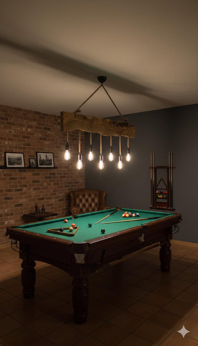 May include: A billiards room featuring a green felt pool table with a wooden frame and legs. Above the table hangs a rustic chandelier with exposed light bulbs. The room has a brick wall and a dark gray wall.