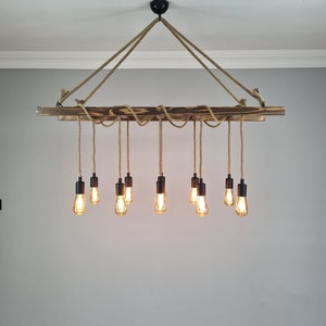 K&ouml;nnte beinhalten: Ein rustikaler Holz-Kronleuchter mit 10 Gl&uuml;hbirnen, die an einem Seil h&auml;ngen. Der Kronleuchter ist aus dunkelbraunem Holz gefertigt und hat ein nat&uuml;rliches Seil-Design.
