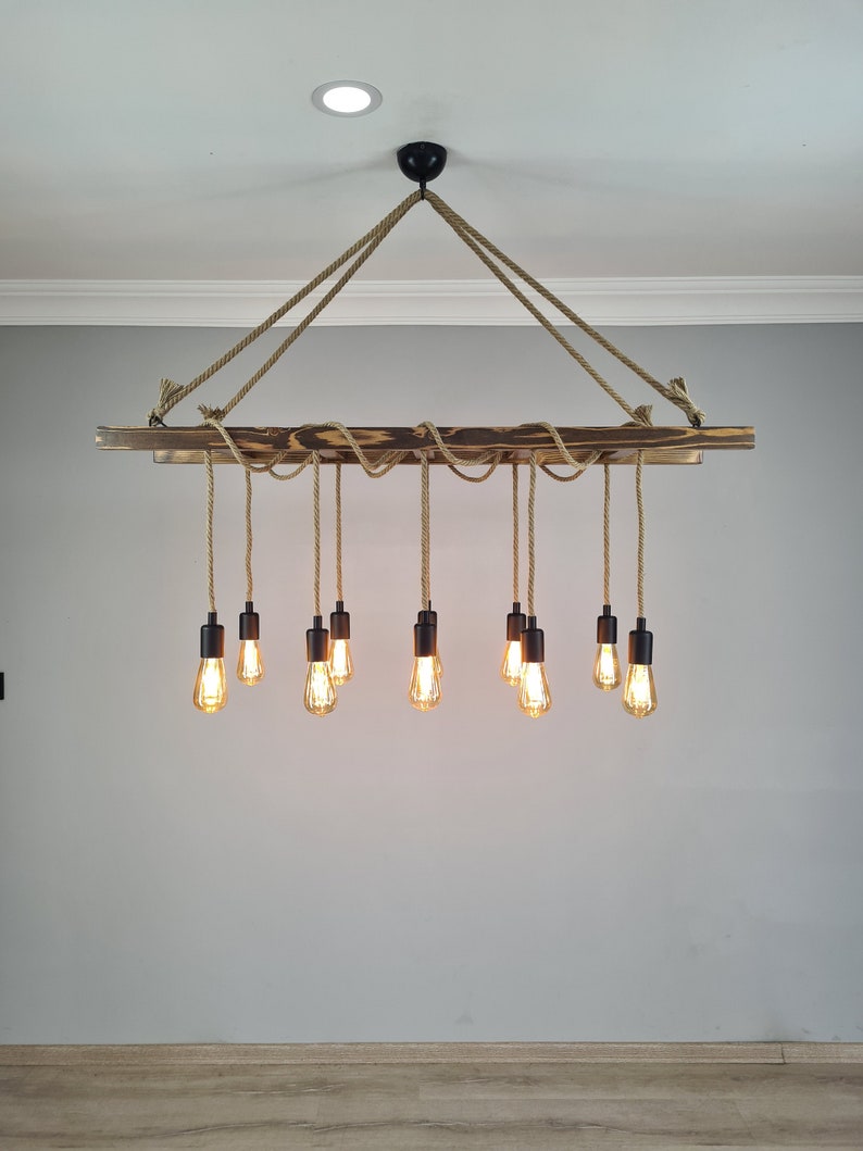 K&ouml;nnte beinhalten: Ein rustikaler Holz-Kronleuchter mit 10 Gl&uuml;hbirnen, die an einem Seil h&auml;ngen. Der Kronleuchter ist aus dunkelbraunem Holz gefertigt und hat ein nat&uuml;rliches Seil-Design.