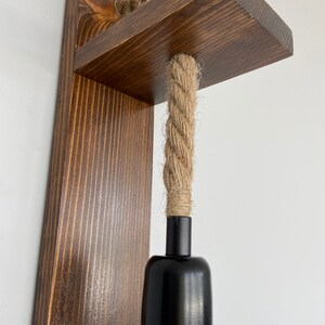 May include: A wall-mounted light fixture featuring a dark brown stained wooden backplate and a horizontal shelf. A thick, natural-colored jute rope passes through the shelf, connecting to a matte black cylindrical light socket hanging below.