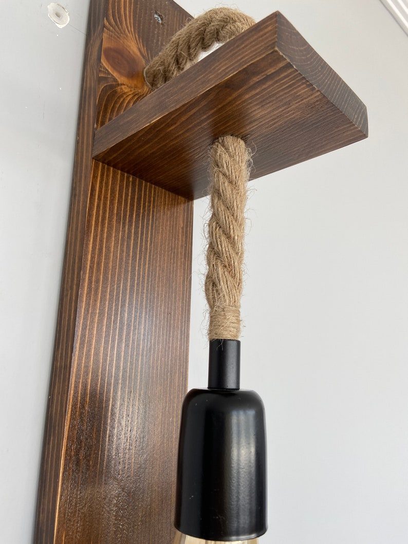 May include: A wall-mounted light fixture featuring a dark brown stained wooden backplate and a horizontal shelf. A thick, natural-colored jute rope passes through the shelf, connecting to a matte black cylindrical light socket hanging below.