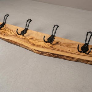 May include: A wooden coat rack with five black metal hooks. The rack is made from a light-colored, live-edge wood, showcasing the natural grain and shape. The hooks are evenly spaced along the length of the wood.