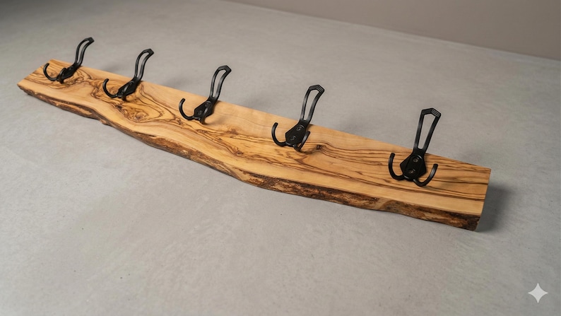 May include: A wooden coat rack with five black metal hooks. The rack is made from a light-colored, live-edge wood, showcasing the natural grain and shape. The hooks are evenly spaced along the length of the wood.