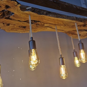 Może przedstawiać: Lampa sufitowa składająca się z dużej drewnianej płyty o naturalnych krawędziach i wyraźnym usłojeniu. Sześć żar&oacute;wek w stylu Edisona z ciemnymi metalowymi oprawkami zwisa na r&oacute;żnych wysokościach, zawieszonych na grubych sznurach w kolorze beżowym.