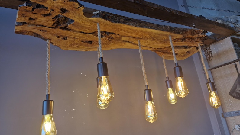 Może przedstawiać: Lampa sufitowa składająca się z dużej drewnianej płyty o naturalnych krawędziach i wyraźnym usłojeniu. Sześć żar&oacute;wek w stylu Edisona z ciemnymi metalowymi oprawkami zwisa na r&oacute;żnych wysokościach, zawieszonych na grubych sznurach w kolorze beżowym.