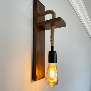 May include: A wall-mounted light fixture featuring a dark stained wooden backplate and a horizontal wooden shelf. A thick, natural-colored jute rope hangs from the shelf, supporting a black socket with a glowing, teardrop-shaped Edison light bulb.