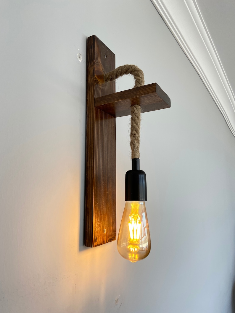 May include: A wall-mounted light fixture featuring a dark stained wooden backplate and a horizontal wooden shelf. A thick, natural-colored jute rope hangs from the shelf, supporting a black socket with a glowing, teardrop-shaped Edison light bulb.