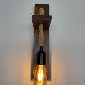 May include: A wall-mounted light fixture featuring a dark brown stained wooden backplate and a horizontal wooden shelf. A thick, natural-colored twisted rope hangs from the shelf, supporting a black socket with a glowing, teardrop-shaped Edison bulb.