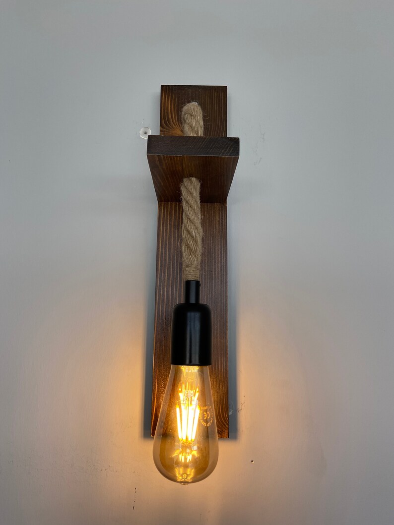 May include: A wall-mounted light fixture featuring a dark brown stained wooden backplate and a horizontal wooden shelf. A thick, natural-colored twisted rope hangs from the shelf, supporting a black socket with a glowing, teardrop-shaped Edison bulb.