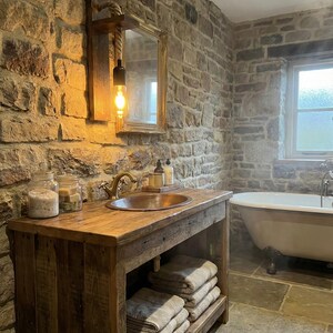 May include: A rustic wooden bathroom vanity featuring a copper sink, a brass faucet, and a lower shelf holding folded towels. Above the vanity, a wall-mounted light fixture with a rope cord and an Edison bulb hangs next to a rectangular mirror.