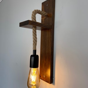 May include: A wall-mounted light fixture featuring a dark brown stained wooden backplate and a small horizontal shelf. A thick, natural-colored jute rope hangs from the shelf, supporting a black socket with a glowing, teardrop-shaped Edison bulb.