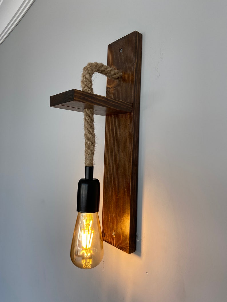 May include: A wall-mounted light fixture featuring a dark brown stained wooden backplate and a small horizontal shelf. A thick, natural-colored jute rope hangs from the shelf, supporting a black socket with a glowing, teardrop-shaped Edison bulb.
