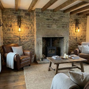 May include: A living room featuring a stone fireplace with a black wood-burning stove, two brown leather armchairs with light gray throw blankets, a wooden coffee table with books and candles, and a large woven rug on a wooden floor.