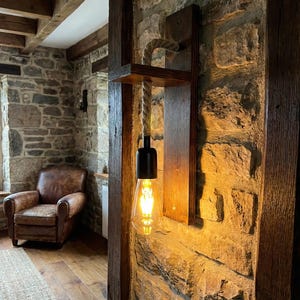 May include: A wall-mounted light fixture featuring a dark brown wooden backplate and a small horizontal shelf. A thick, natural-colored rope hangs from the shelf, supporting a black socket with a glowing, teardrop-shaped Edison-style light bulb.