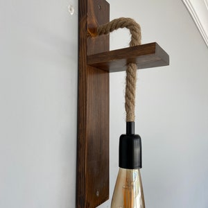 May include: A wall-mounted light fixture featuring a dark brown stained wooden backplate and a horizontal wooden shelf. A thick, natural-colored jute rope passes through the shelf, connecting to a black socket holding a clear, teardrop-shaped bulb.