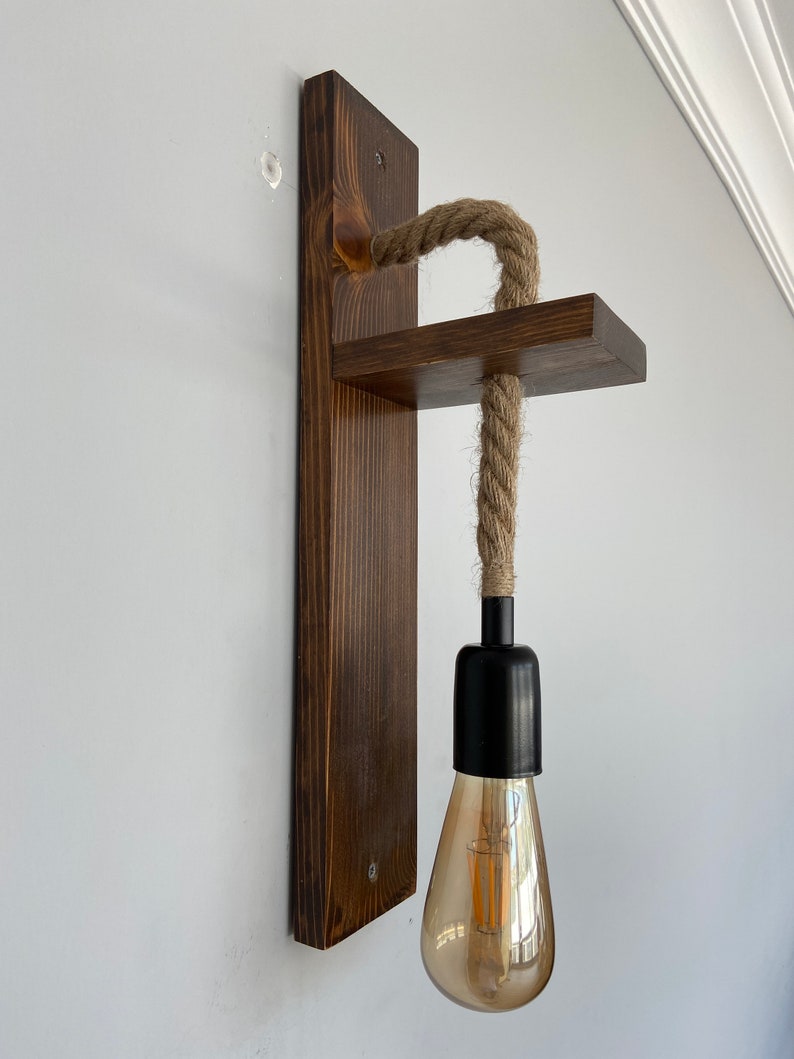 May include: A wall-mounted light fixture featuring a dark brown stained wooden backplate and a horizontal wooden shelf. A thick, natural-colored jute rope passes through the shelf, connecting to a black socket holding a clear, teardrop-shaped bulb.
