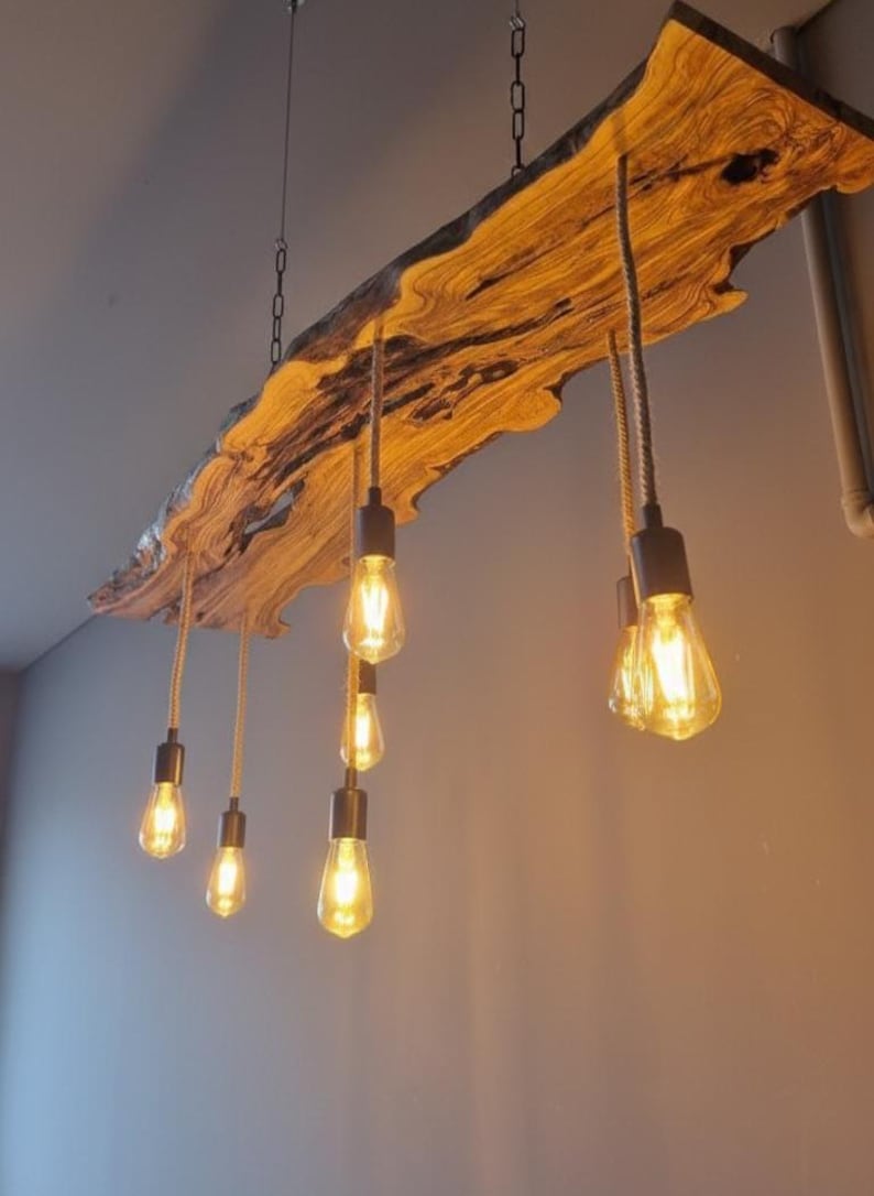 Może przedstawiać: Lampa sufitowa wykonana z drewnianej płyty z naturalną krawędzią, zawieszona na metalowych łańcuchach. Oprawa posiada sześć zwisających żar&oacute;wek z ciepło świecącymi żarnikami, przymocowanych do drewna za pomocą grubych sznur&oacute;w i ciemnych oprawek.