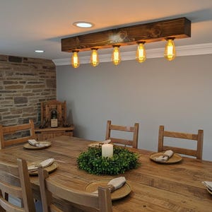 May include: A rectangular dark stained wooden beam ceiling light fixture featuring five hanging Edison-style light bulbs with warm glowing filaments, suspended horizontally above a wooden dining table set with plates and napkins.