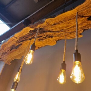 Może przedstawiać: Rustykalna lampa wisząca z długą, naturalną deską z widocznym usłojeniem. Pięć żar&oacute;wek w stylu Edisona zwisa z drewna na grubych, teksturowanych sznurach, każda zamocowana w matowej, czarnej metalowej oprawce.