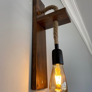 May include: A wall-mounted light fixture featuring a dark stained wooden backplate and a horizontal wooden arm. A thick, natural-colored jute rope hangs from the arm, supporting a black socket with a glowing, teardrop-shaped Edison light bulb.